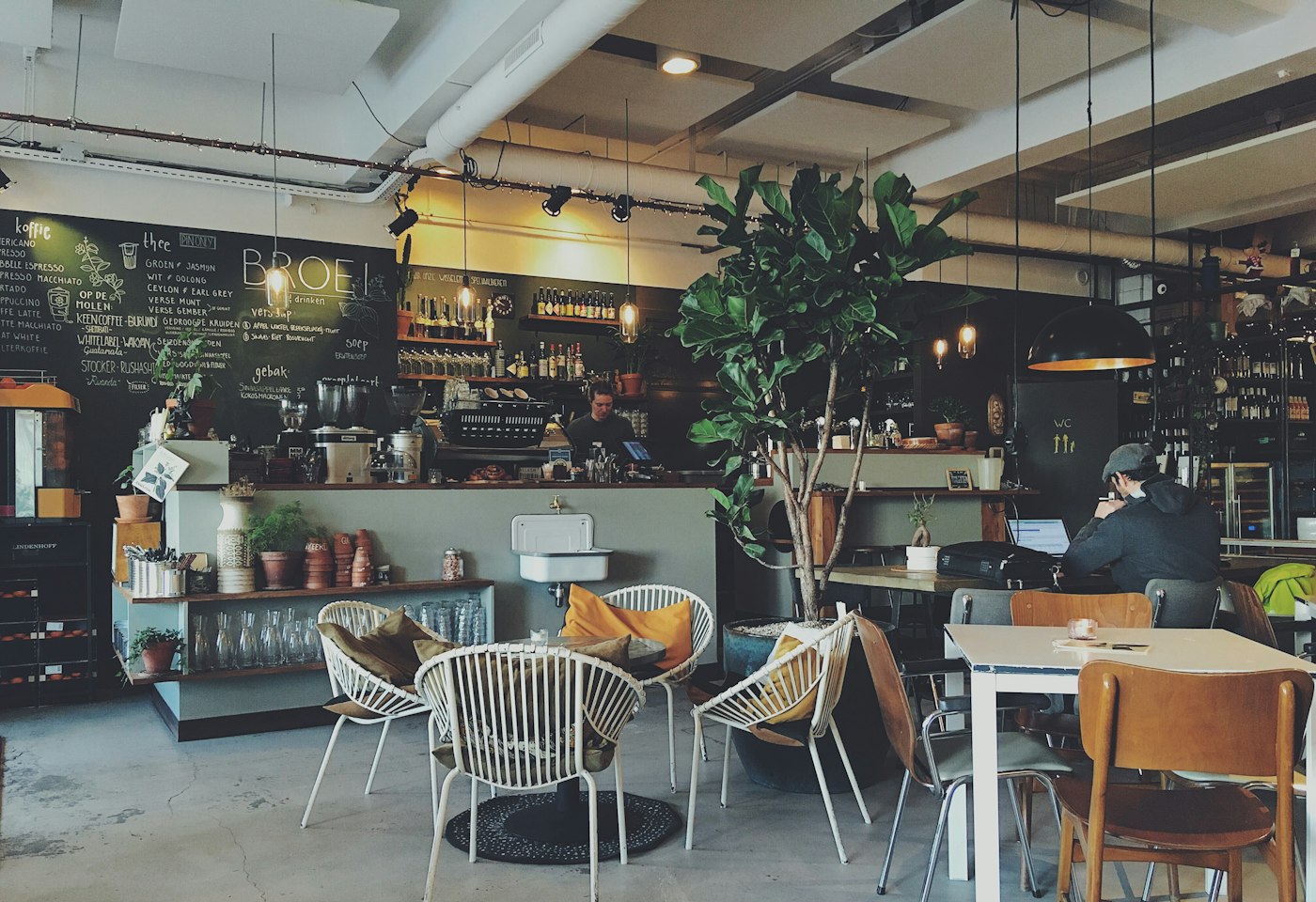 Café interior with a coffee counter, chalkboard menu, plants, and guests at tables